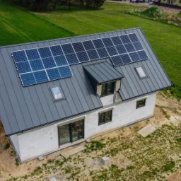 Widok z góry na dom w trakcie budowy z zamontowanymi panelami słonecznymi na szarym dachu z blachy trapezowej, okna dachowe i lukarna, otoczony zielonym trawnikiem i ziemią.