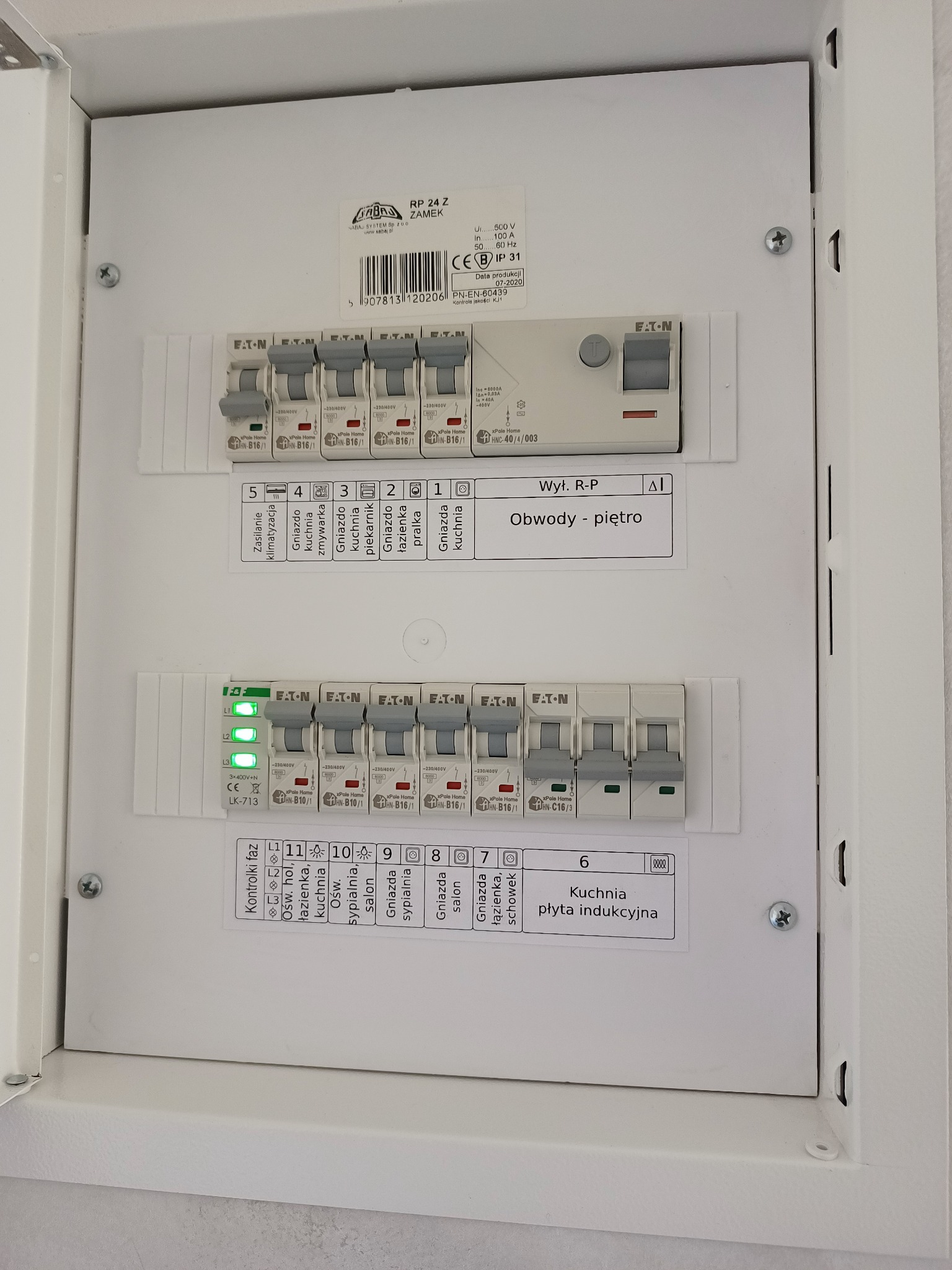 Biała skrzynka elektryczna z bezpiecznikami, opisane obwody, widoczne kontrolki faz i etykiety z opisem gniazd i urządzeń, w tym płyty indukcyjnej w kuchni i zasilania klimatyzacji.