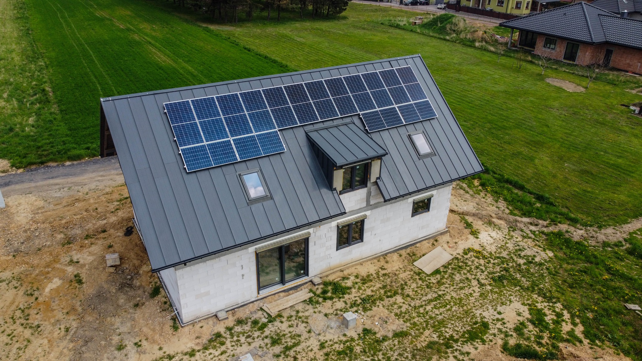 Widok z góry na dom w trakcie budowy z zamontowanymi panelami słonecznymi na szarym dachu z blachy trapezowej, okna dachowe i lukarna, otoczony zielonym trawnikiem i ziemią.