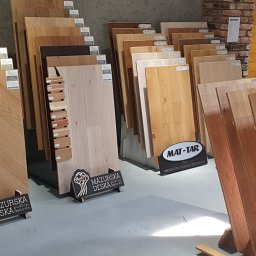 Fabryka Podłogi Drzwi - Ekspozycja różnych rodzajów podłóg drewnianych, w tym deski dębowe w jodełkę, z widocznymi próbkami kolorystycznymi i oznaczeniami producentów Mazurska Deska i Mat-Tar.