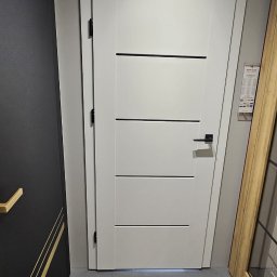 Fabryka Podłogi Drzwi - Białe drzwi wewnętrzne marki Erkado z trzema poziomymi, czarnymi wstawkami, prezentowane w salonie obok innych modeli.