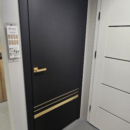 Fabryka Podłogi Drzwi - Czarne drzwi wewnętrzne Erkado Altamura z złotymi akcentami, prezentowane w salonie obok białych drzwi. Widoczny katalog z wzorami i logo producenta.