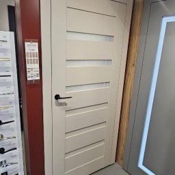 Fabryka Podłogi Drzwi - Beżowe drzwi wewnętrzne z poziomymi przeszkleniami, czarna klamka, ekspozycja w salonie, obok katalog z okuciami i inne drzwi.