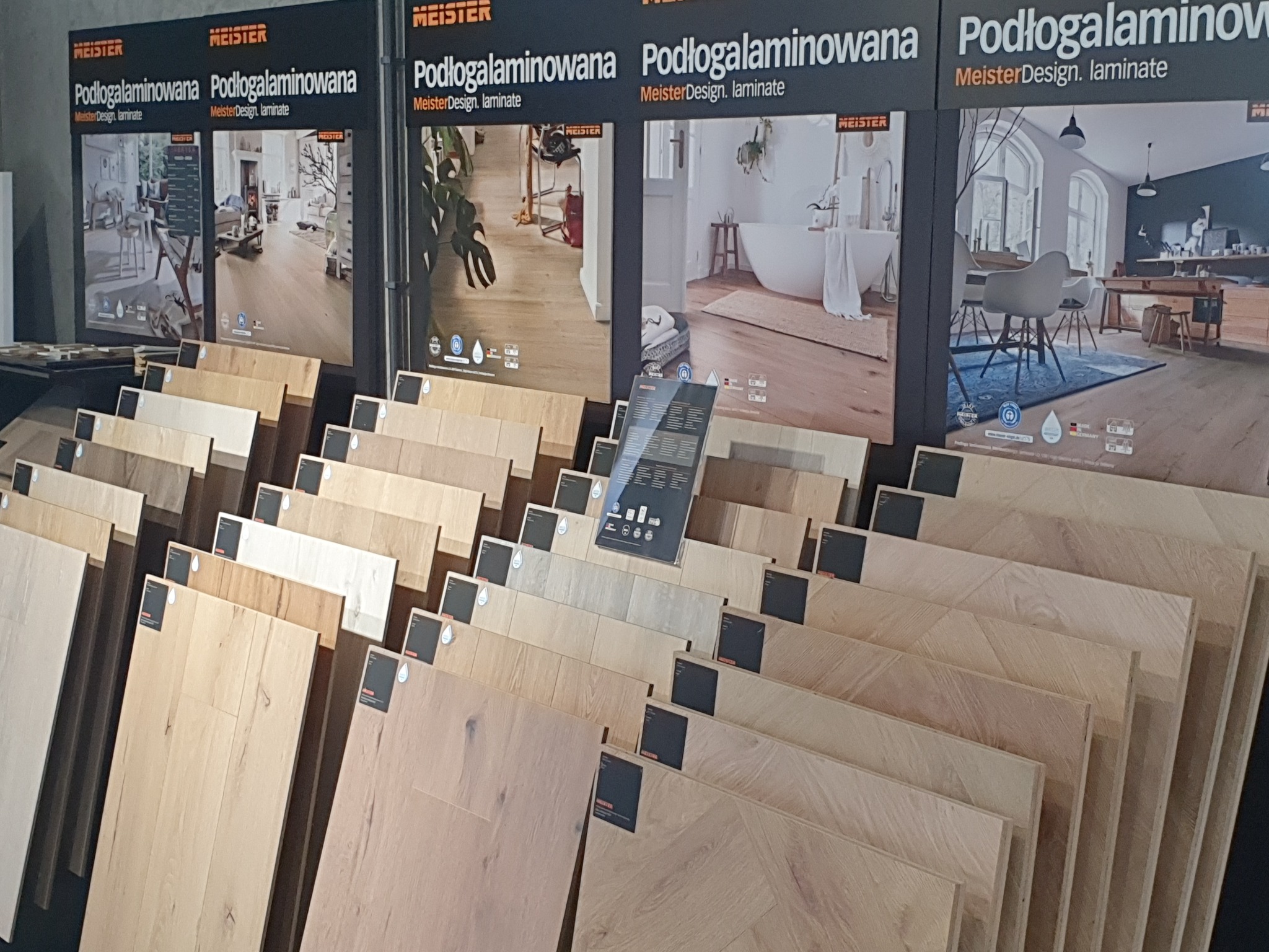 Ekspozycja próbek laminowanych paneli podłogowych w różnych odcieniach drewna, z plakatami reklamowymi producenta w tle prezentującymi aranżacje wnętrz z ich użyciem.