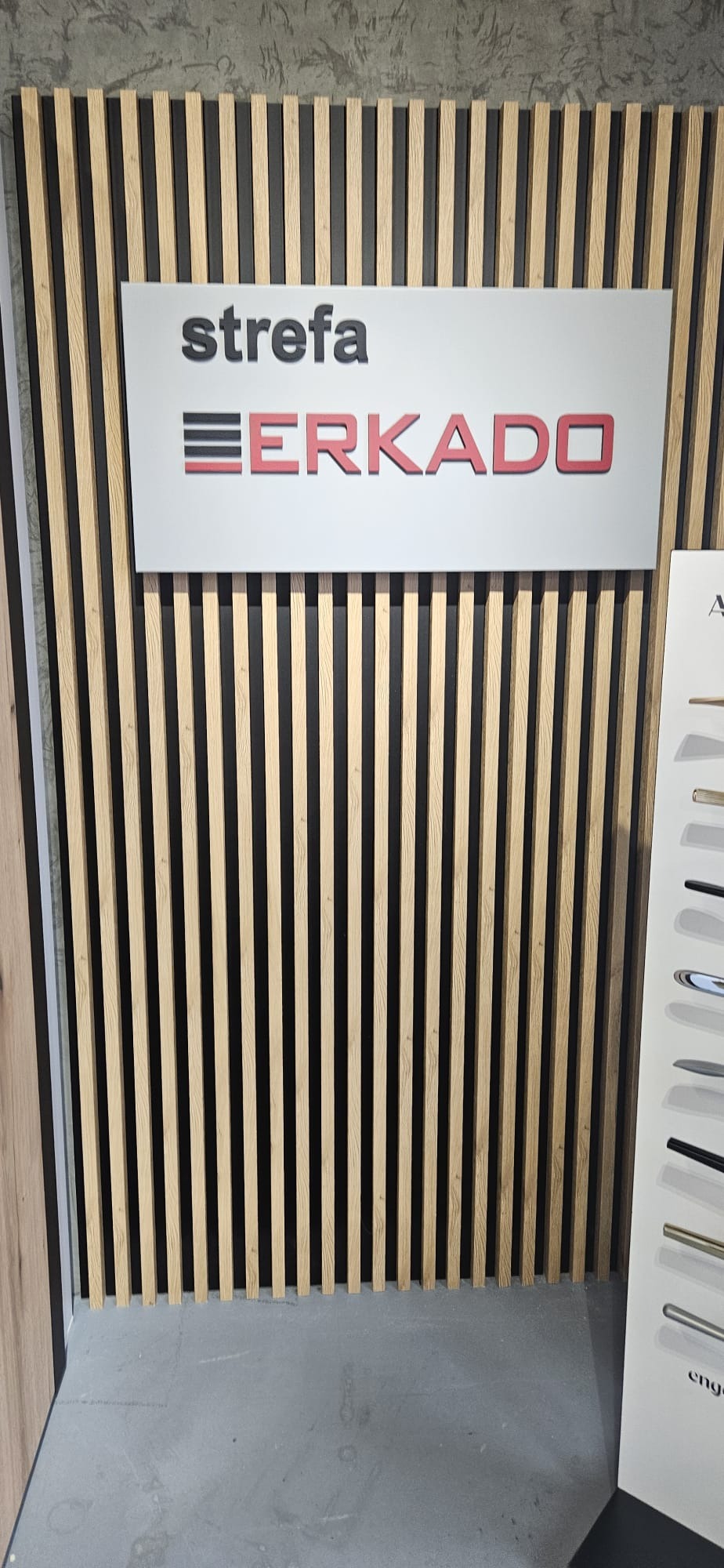 Drewniane lamele na czarnym tle, z logotypem 'Strefa ERKADO' na białej tablicy. Po prawej ekspozycja klamek. Dekoracja wnętrza w nowoczesnym stylu.