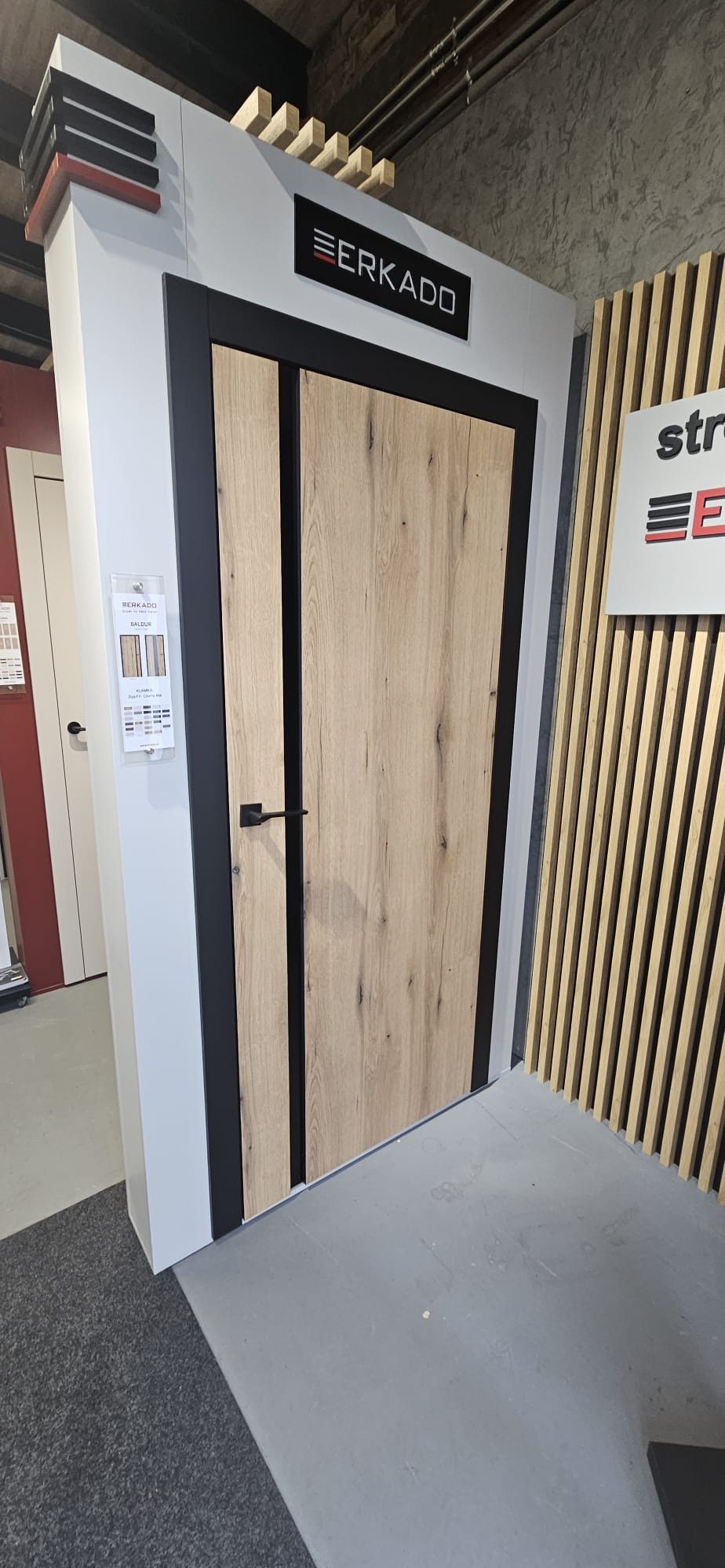 Drewnopodobne drzwi Erkado z czarną ramą, prezentowane w salonie. Widoczne detale wykończenia i ekspozycja z katalogiem.