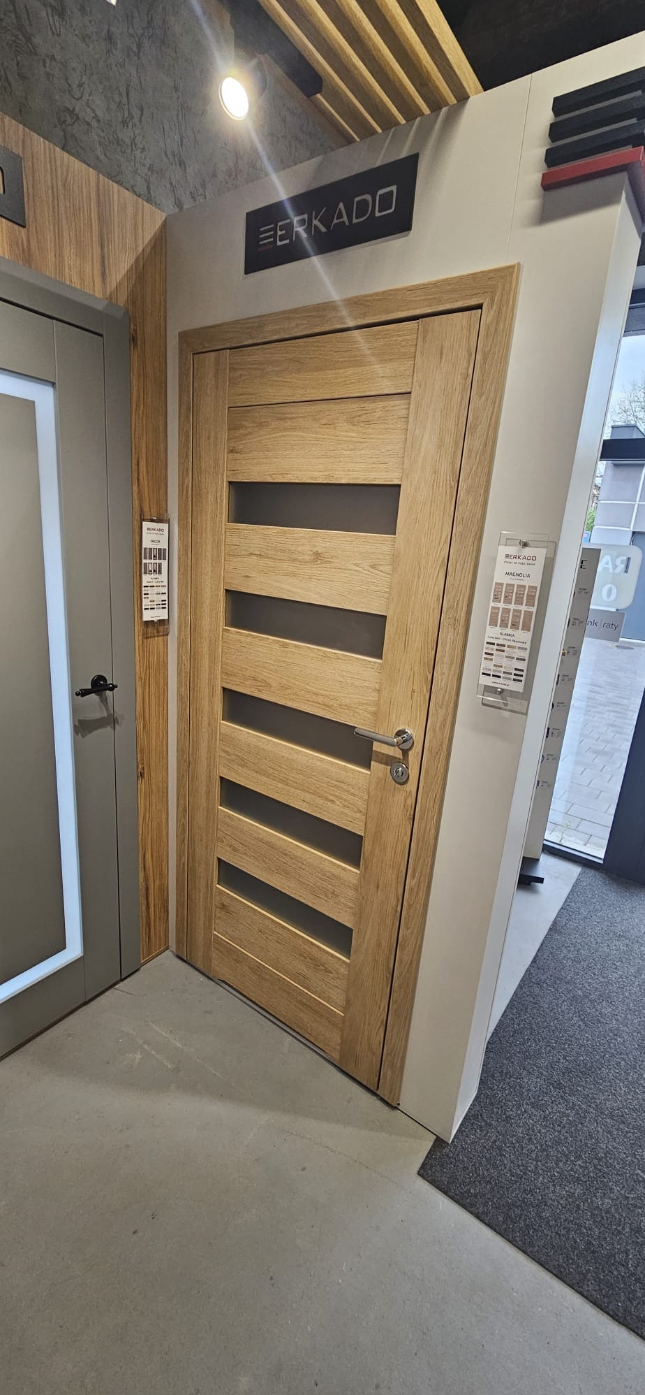 Jasne, drewniane drzwi wewnętrzne z czterema poziomymi przeszkleniami, prezentowane w salonie ekspozycyjnym marki Erkado. Obok drzwi katalog z wyborem kolorów.
