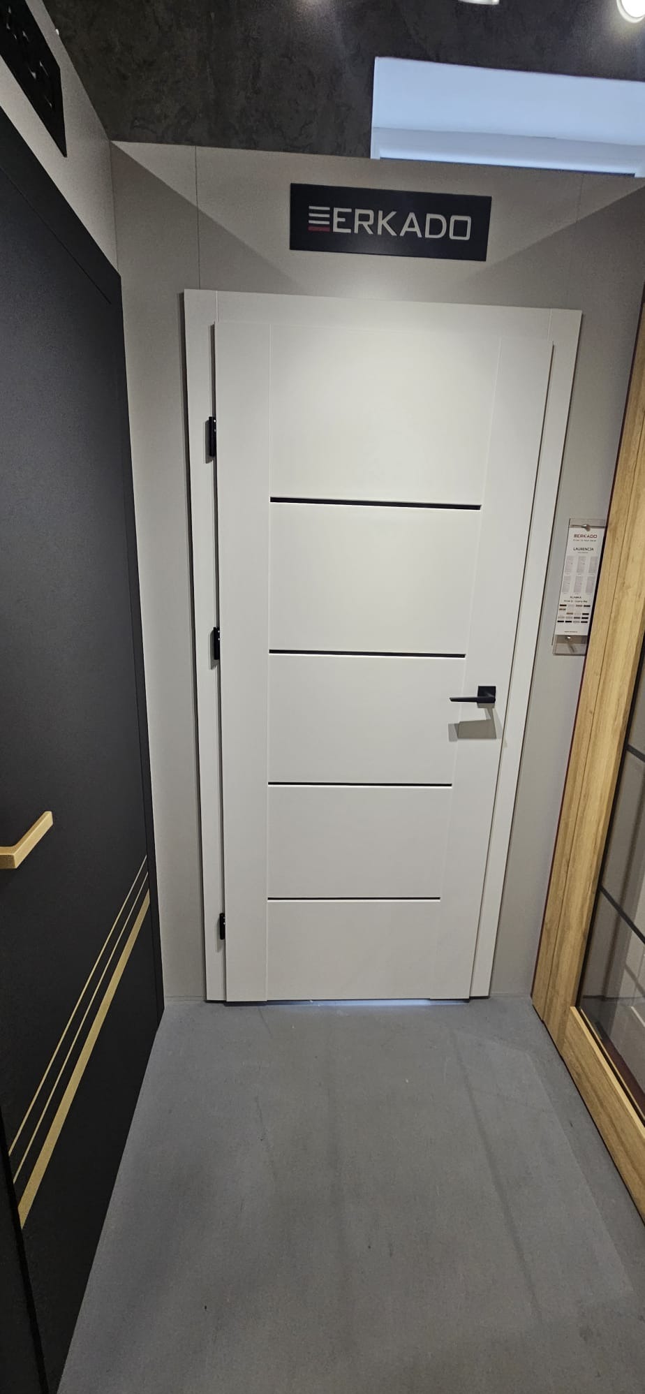 Białe drzwi wewnętrzne marki Erkado z trzema poziomymi, czarnymi wstawkami, prezentowane w salonie obok innych modeli.