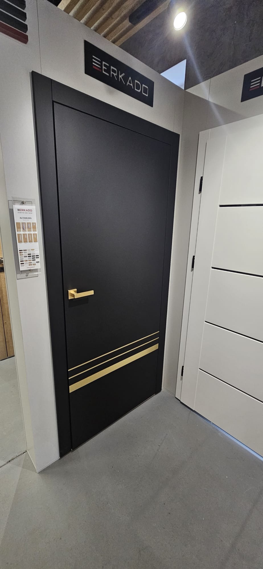 Czarne drzwi wewnętrzne Erkado Altamura z złotymi akcentami, prezentowane w salonie obok białych drzwi. Widoczny katalog z wzorami i logo producenta.