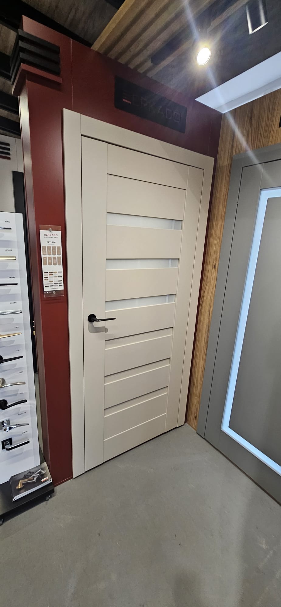 Beżowe drzwi wewnętrzne z poziomymi przeszkleniami, czarna klamka, ekspozycja w salonie, obok katalog z okuciami i inne drzwi.