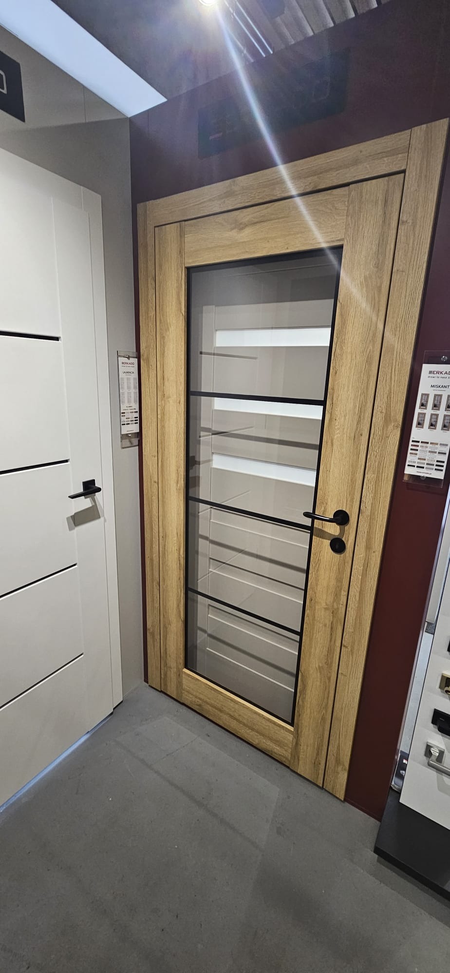 Drewniane drzwi wewnętrzne z szybą i czarnymi szprosami, widoczne obok białe drzwi z poziomymi frezami, prezentowane w salonie sprzedaży.