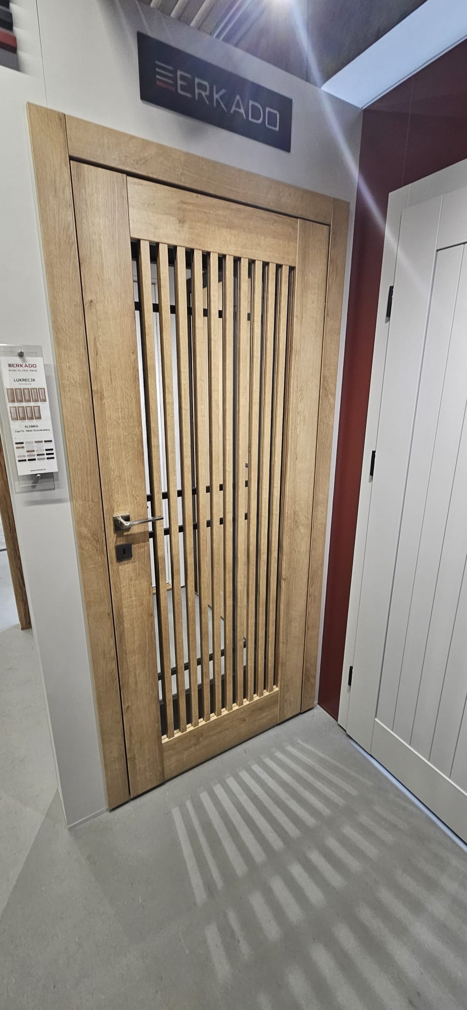 Drewniane drzwi wewnętrzne z pionowymi szczebelkami i metalową klamką na tle ekspozycji z logo marki Erkado, obok białe drzwi.