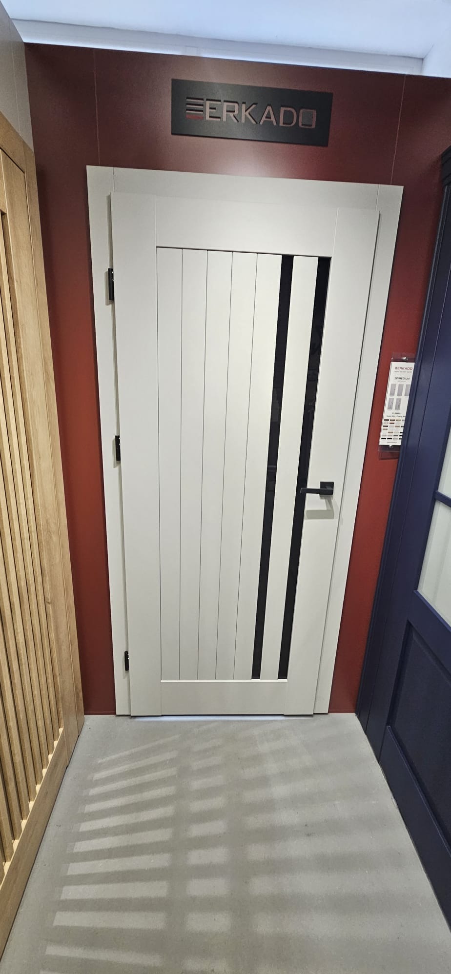 Białe drzwi wewnętrzne z czarnymi, pionowymi wstawkami, prezentowane w salonie. Widoczne logo producenta 'Eerkado' i fragmenty innych modeli drzwi.