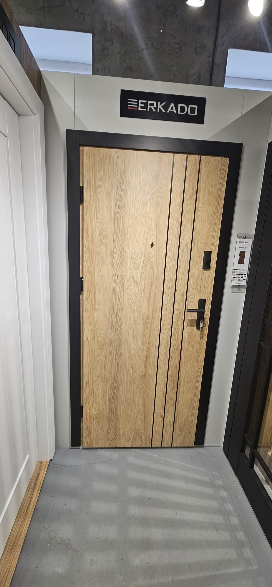 Drewniane drzwi Erkado z czarną ramą, z delikatnymi, pionowymi wstawkami. Prezentacja w salonie, widoczny fragment podłogi i sąsiednich drzwi.