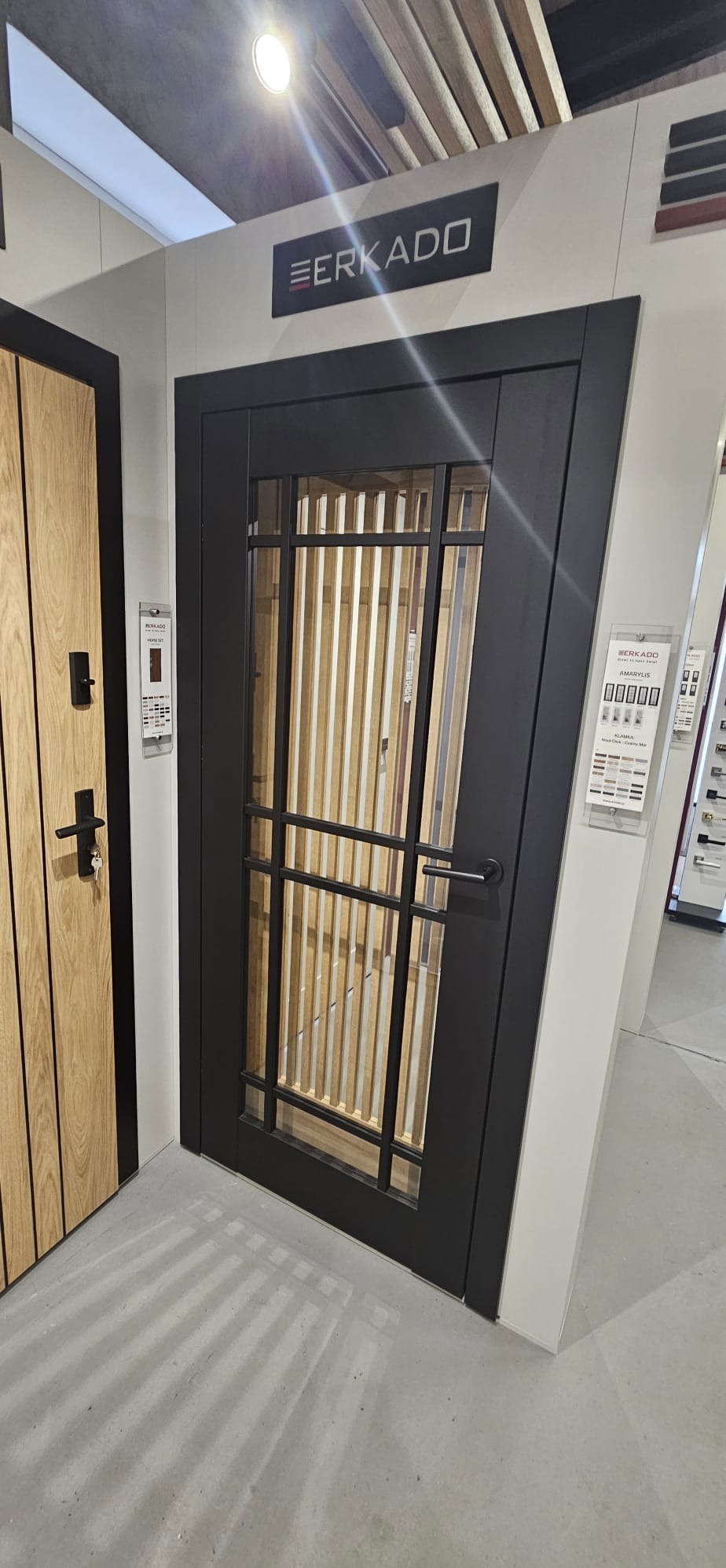 Czarne drzwi wewnętrzne z przeszkleniami w stylu loft, widoczne na ekspozycji w salonie, obok drzwi z drewnianą okleiną i czarnym zamkiem.