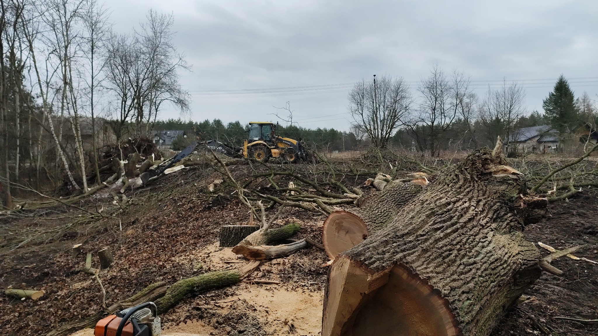 Żółta koparko-ładowarka Volvo na skarpie z powalonymi drzewami i gałęziami, w tle budynek mieszkalny, piła spalinowa na pierwszym planie, pochmurne niebo.