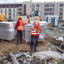 Trater - Pracownicy w kaskach i kamizelkach transportują izolowane panele na placu budowy. W tle budynki mieszkalne i przenośne toalety.