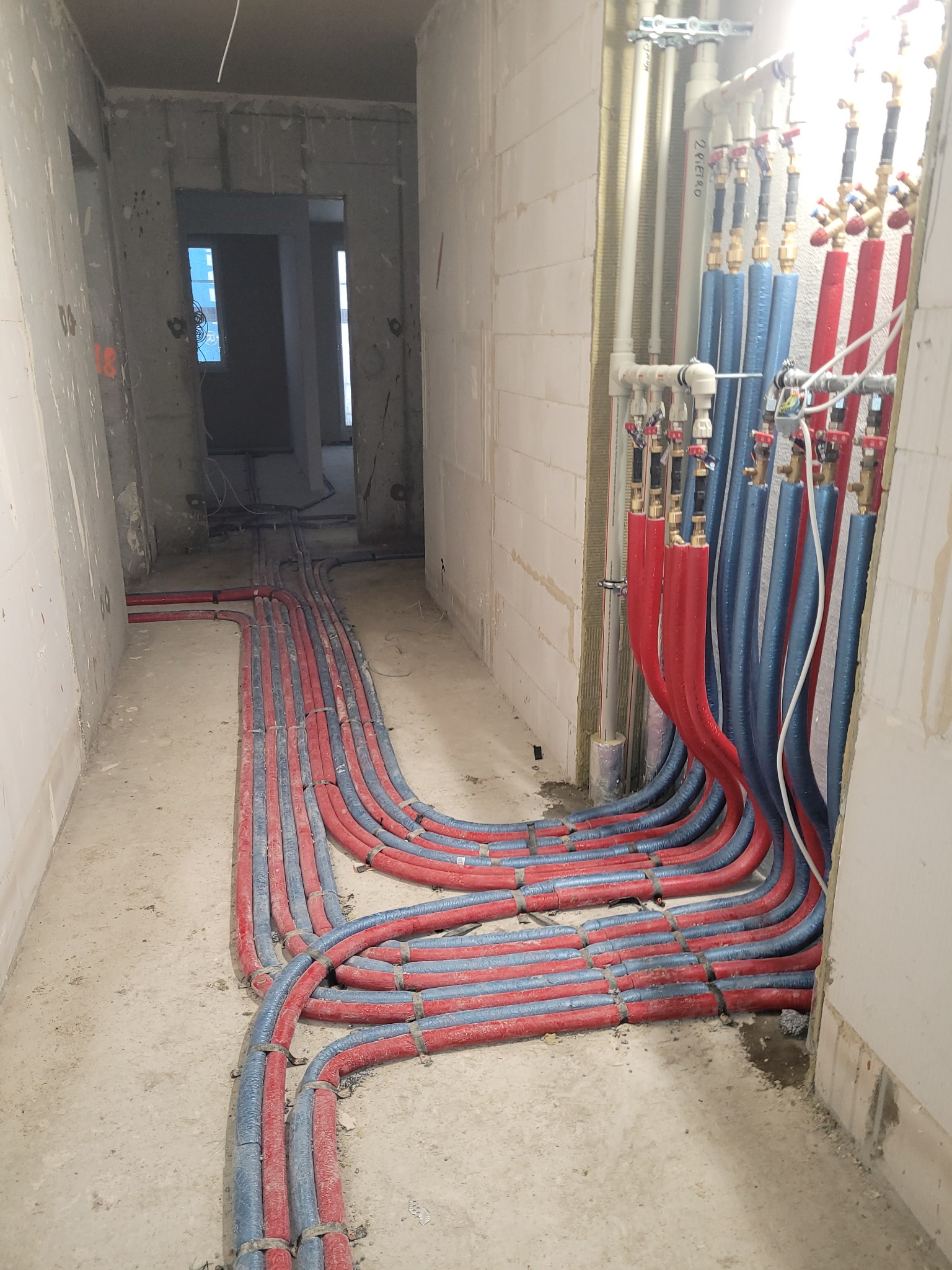 Instalacja rur w budynku w stanie surowym: czerwone i niebieskie rury biegną wzdłuż podłogi i ściany, prowadząc do pionowych rur z zaworami. Widok z perspektywy.