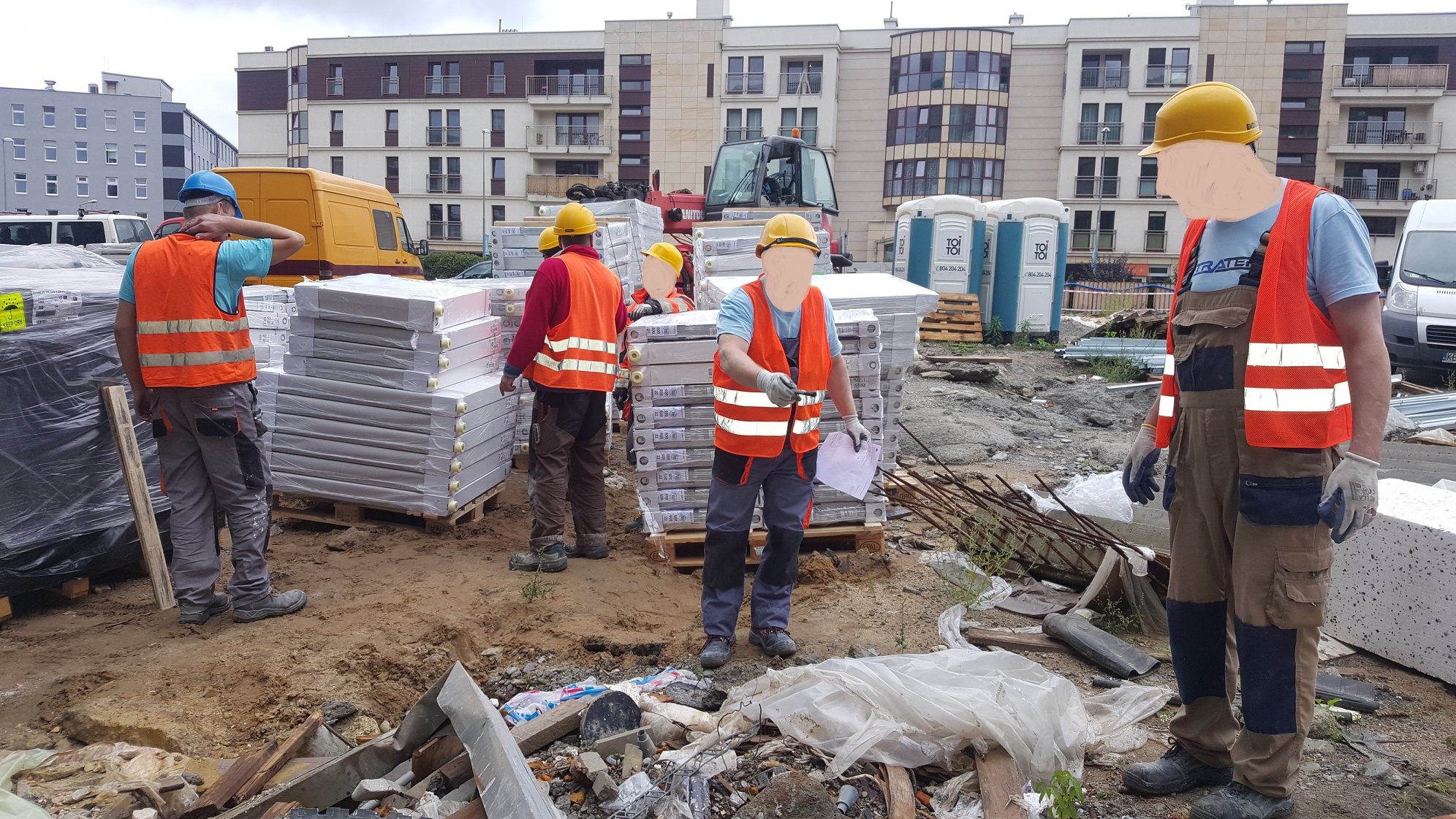 Pracownicy w kaskach i kamizelkach transportują izolowane panele na placu budowy. W tle budynki mieszkalne i przenośne toalety.