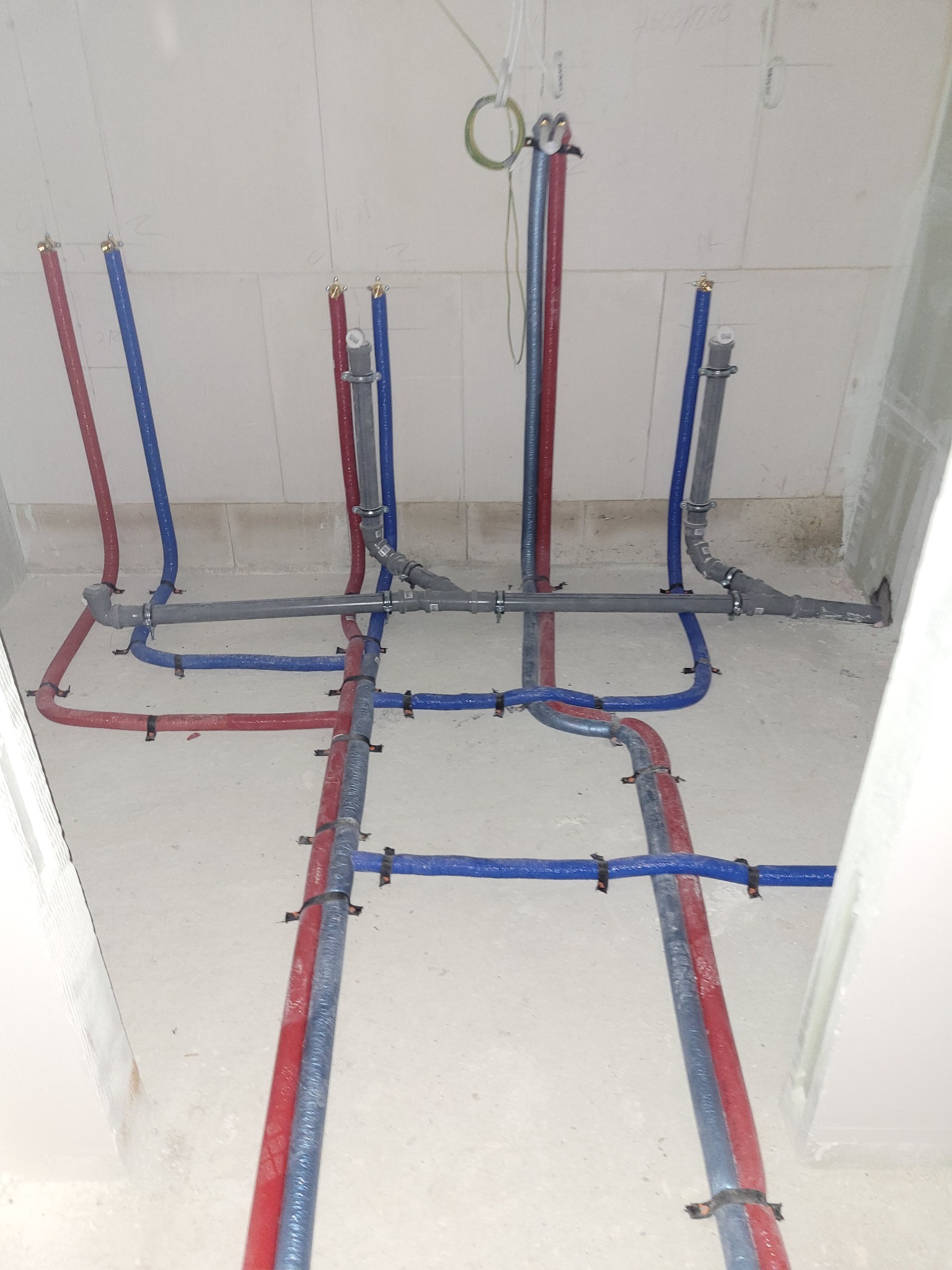 Rozprowadzenie instalacji wodnej z rur PEX w kolorze niebieskim i czerwonym, przymocowanych do podłoża. Szare rury kanalizacyjne złączone złączkami.