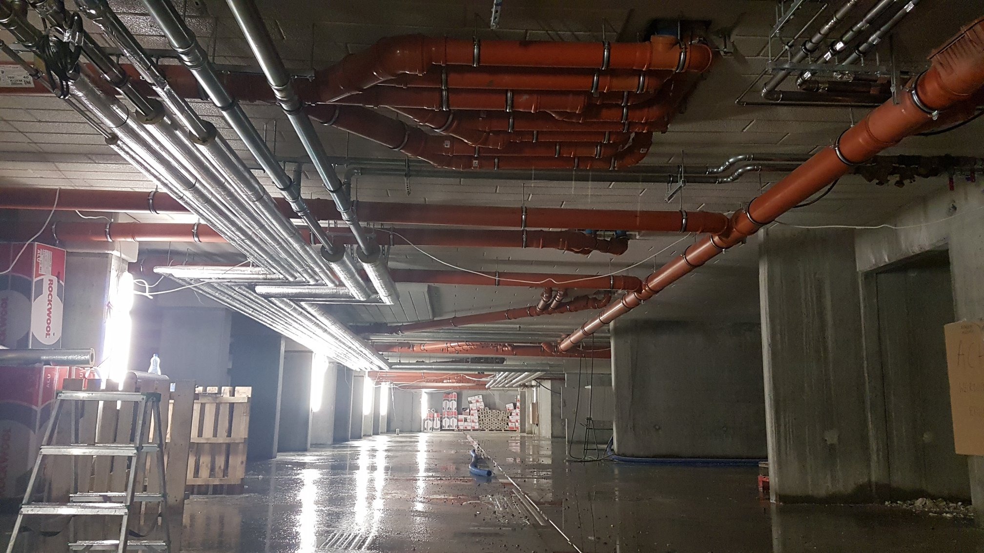 Rozbudowana instalacja sanitarna z rur PCV i metalowych, zamontowana pod stropem surowego budynku w trakcie budowy. Widoczne mocowania i elementy podwieszenia.