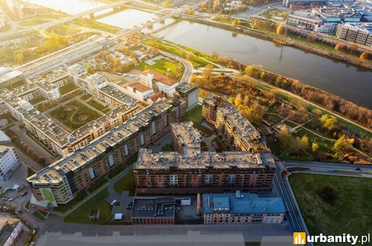 Widok z lotu ptaka na nowoczesne osiedle budynków mieszkalnych z ceglanymi elewacjami, otoczone zielenią i rzeką, oświetlone ciepłym, popołudniowym światłem.