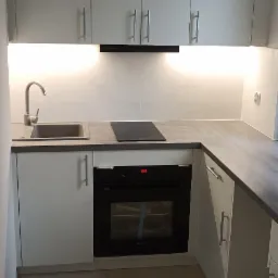 Nowoczesna kuchnia w odcieniach bieli i szarości z oświetleniem podszafkowym, widok na zlewozmywak, płytę indukcyjną i piekarnik z folią ochronną na drzwiczkach.