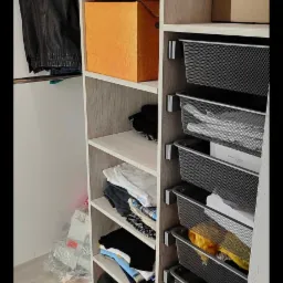 Wnętrze otwartej szafy z jasnego drewna z półkami i metalowymi koszami wypełnionymi ubraniami, torbą i pudełkami.