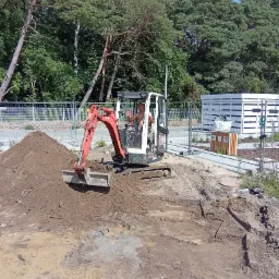 Mini koparka Kubota w trakcie prac ziemnych na terenie zielonym z drzewami w tle, operator w kabinie, widoczne ślady opon i przesypana ziemia.