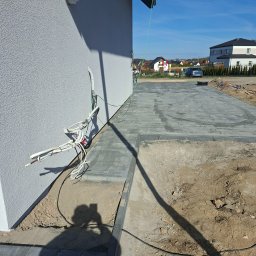 Arko-Bud - Układanie szarej kostki brukowej na podjeździe w Elblągu. Widoczne wystające kable elektryczne przy ścianie budynku i cień osoby robiącej zdjęcie.