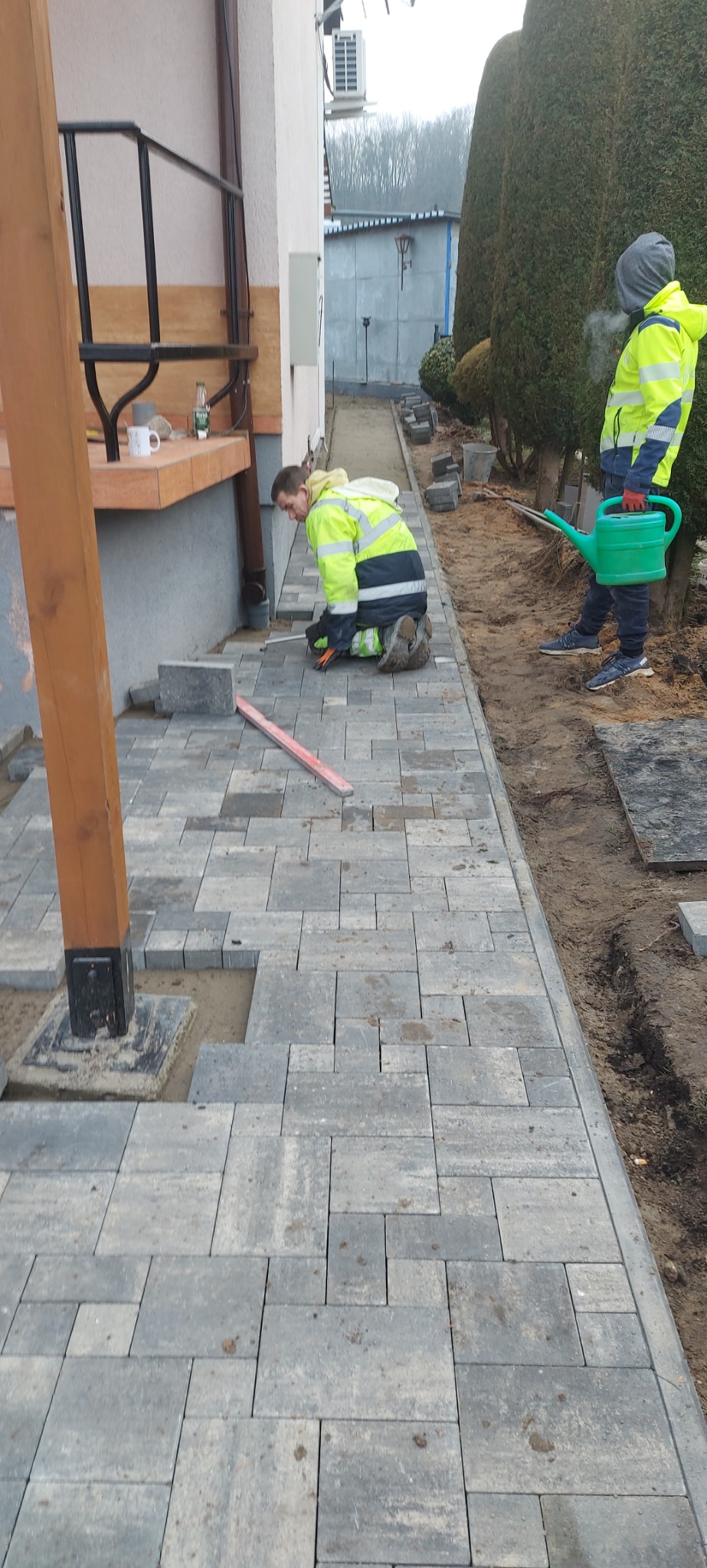 Pracownik w jaskrawej kurtce układa szare kostki brukowe na podwórku domu jednorodzinnego, drugi podlewa teren zielony obok, widoczny poziomica.