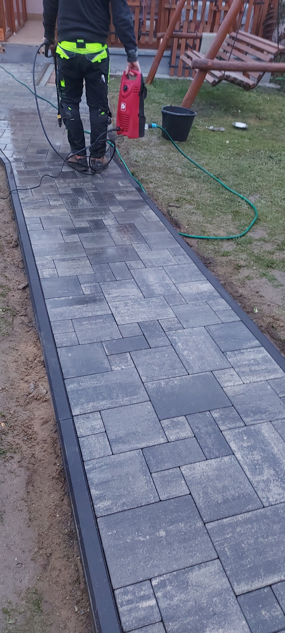 Umyta część ścieżki z kostki brukowej w odcieniach szarości, obramowana czarnym obrzeżem, z pracownikiem w odzieży roboczej czyszczącym ją myjką ciśnieniową. W tle huśtawka ogrodowa i trawnik.