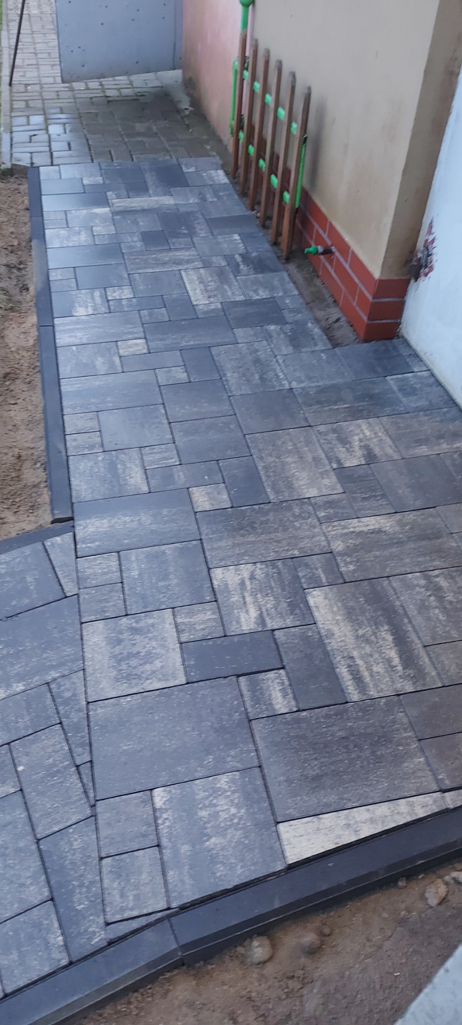 Ułożona ścieżka z szarej kostki brukowej o nieregularnym wzorze, obrzeża z czarnej kostki, widok z góry, obok ściana budynku z widocznymi cegłami i elementami instalacji.