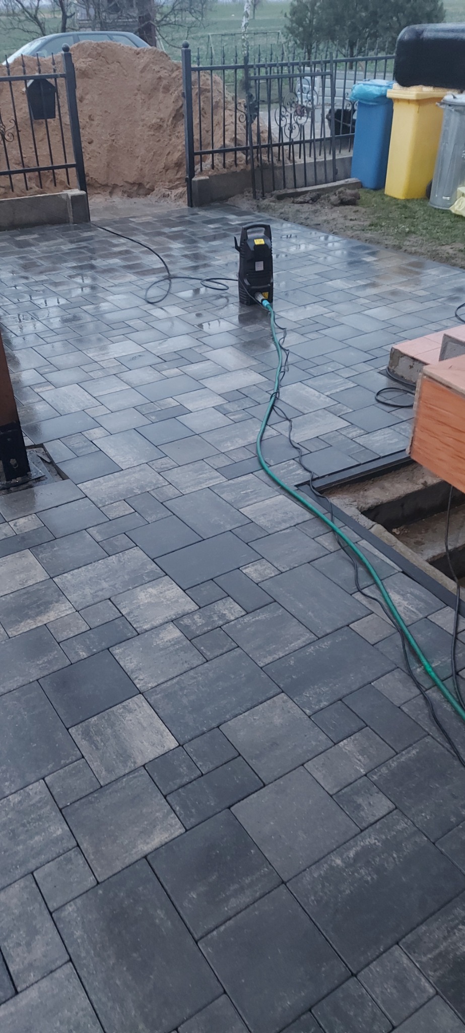 Świeżo ułożona, mokra kostka brukowa w odcieniach szarości, czyszczona myjką ciśnieniową. Widoczne przygotowania do dalszych prac wykończeniowych.