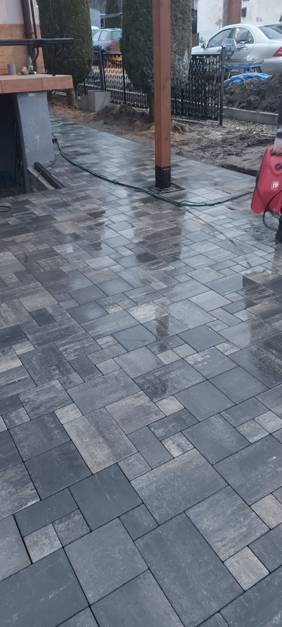 Świeżo ułożona, mokra kostka brukowa w odcieniach szarości tworzy wzór na podjeździe, widoczny fragment drewnianej konstrukcji i czarny wąż ogrodowy.