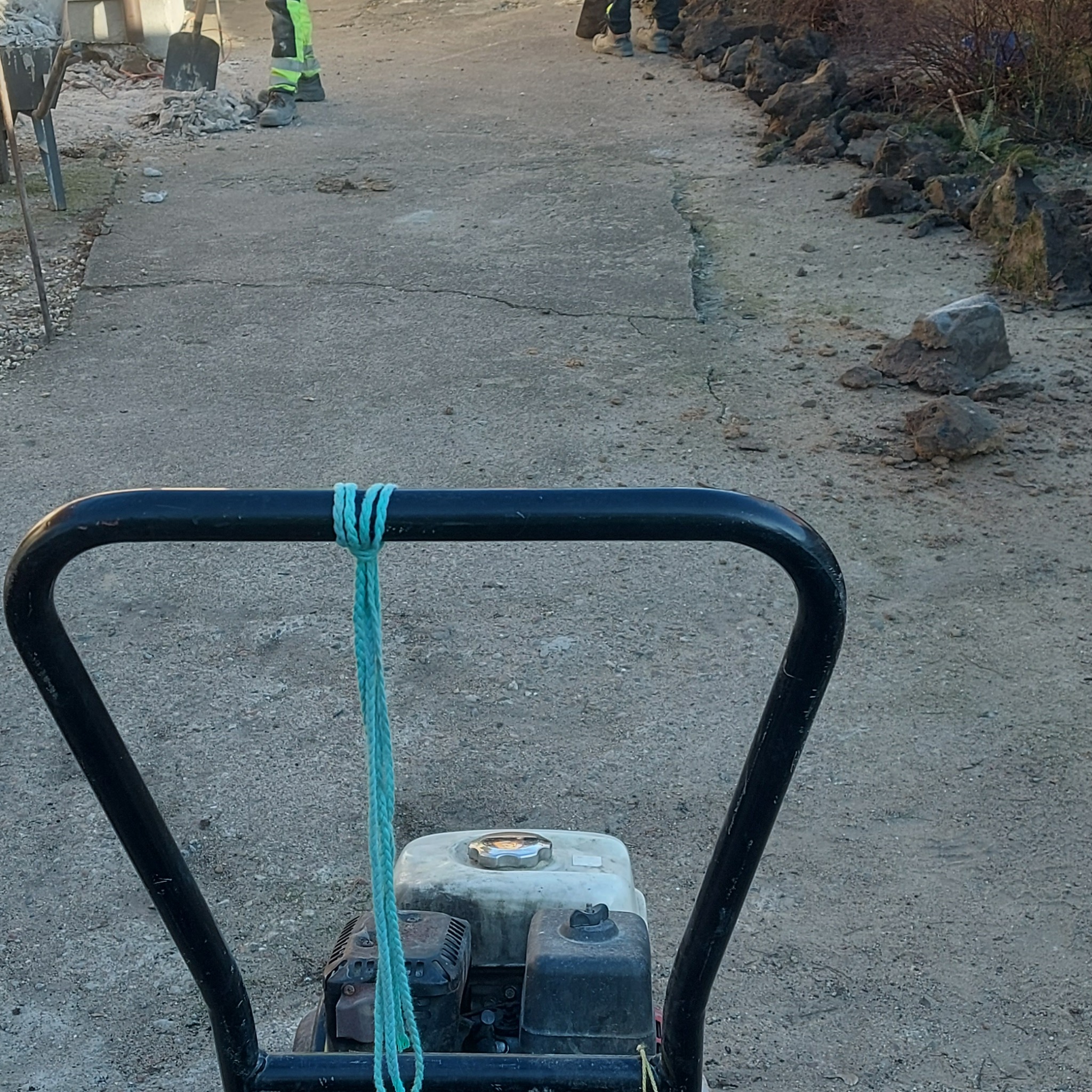 Praca brukarska w Elblągu: Zagęszczarka wibracyjna na tle przygotowywanej powierzchni, w tle pracownicy z łopatami i pryzma gruzu.