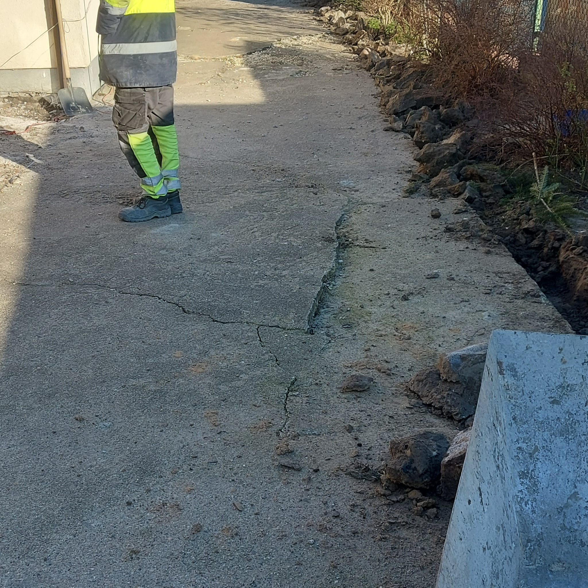 Pęknięta betonowa ścieżka przed domem, przygotowana do naprawy lub wymiany, z widocznym fragmentem pracownika w odblaskowej kamizelce i jaskrawych spodniach oraz odsłoniętym gruntem po bokach.