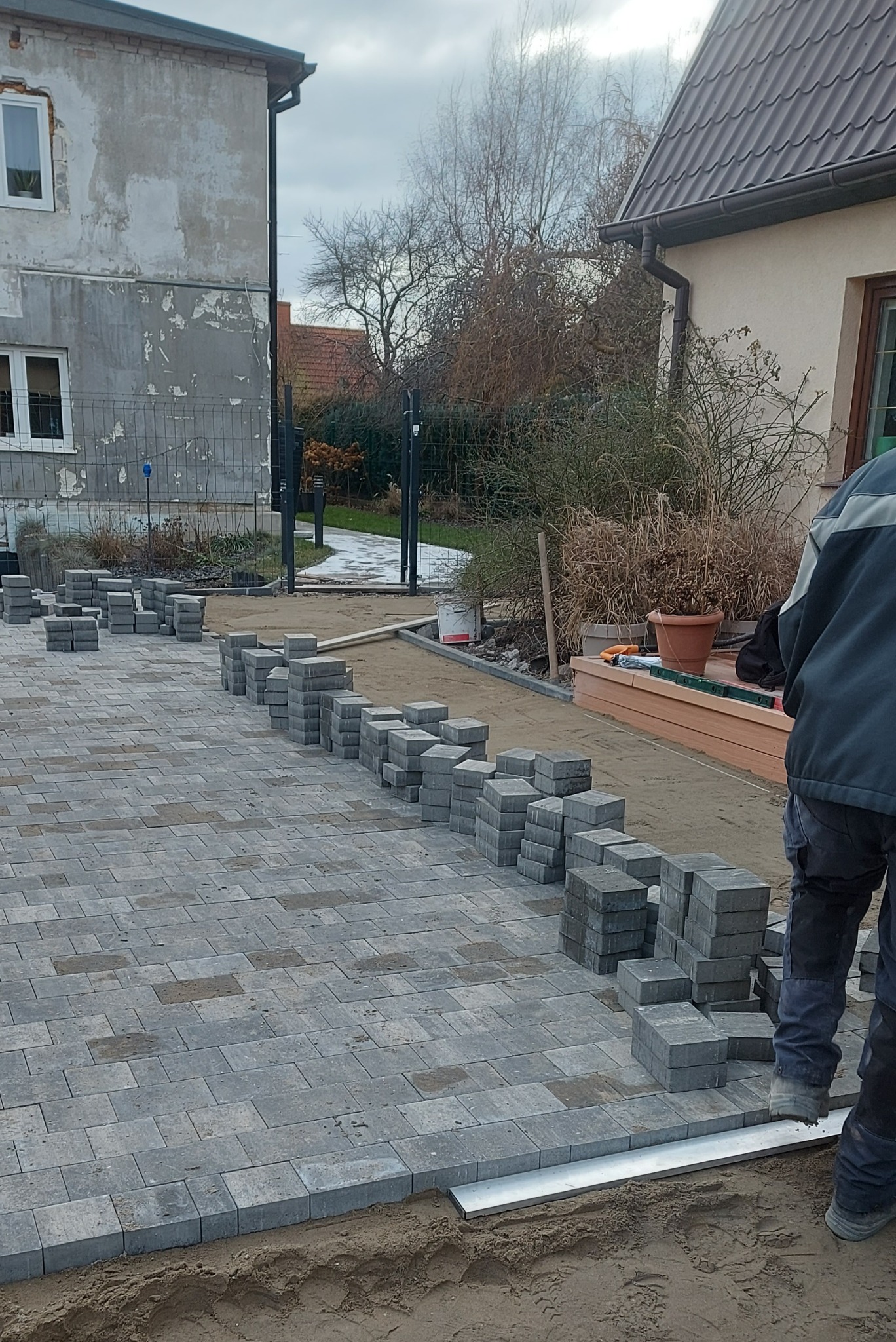 Układanie szarej kostki brukowej na podwórku w Elblągu, widoczne stosy kostki przygotowane do ułożenia, częściowo ułożona nawierzchnia z kostki.