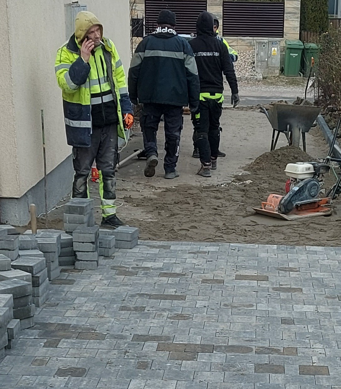 Pracownicy w odzieży roboczej układają szarą kostkę brukową na podwórku, jeden z nich rozmawia przez telefon, w tle ubijarka i taczka.