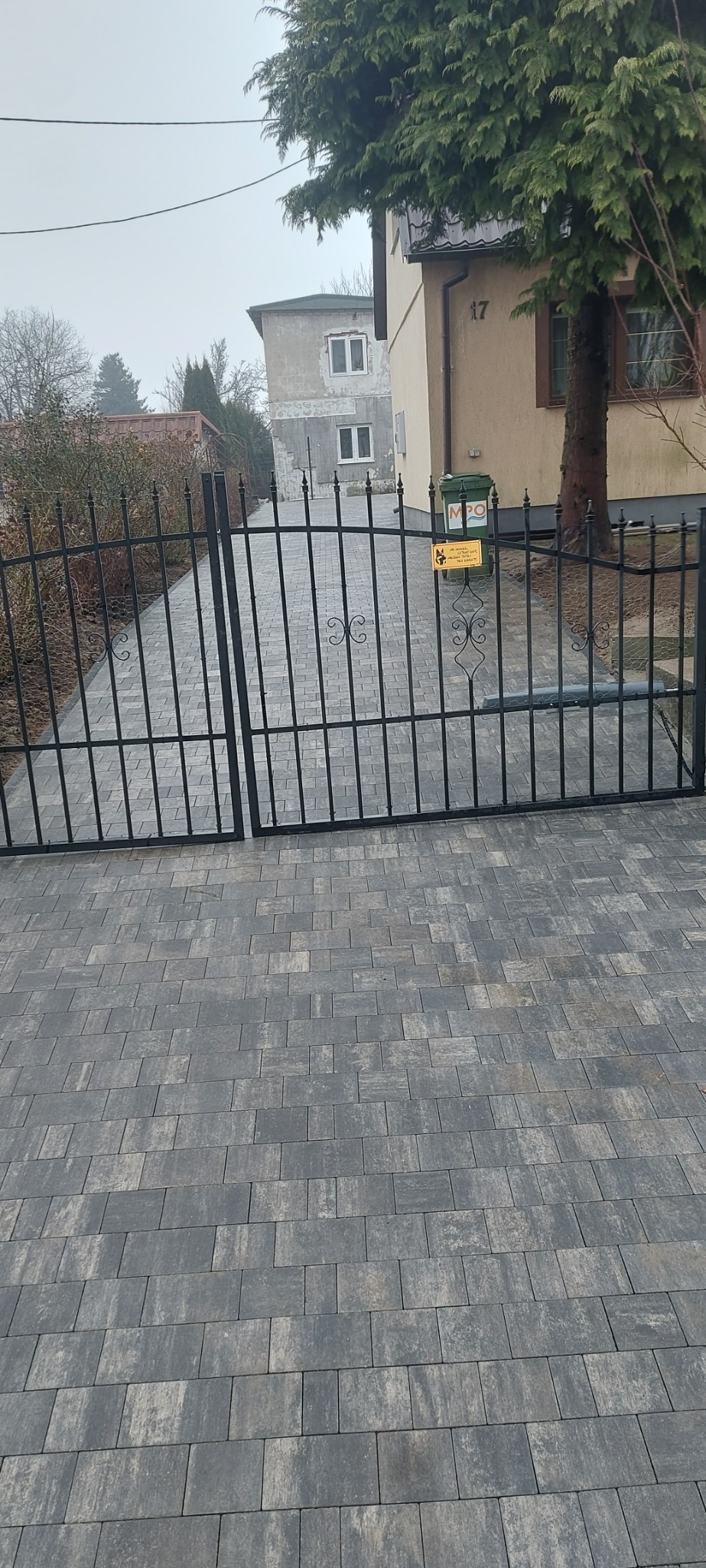 Nowo ułożona szara kostka brukowa prowadząca do czarnej, metalowej bramy wjazdowej, z domem w tle w pochmurny dzień.