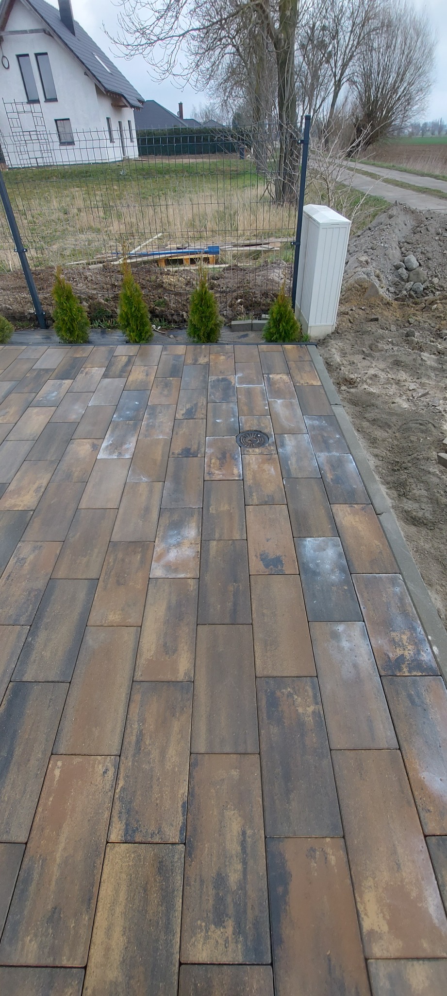Nowa nawierzchnia z kostki brukowej o odcieniach brązu i szarości, z małą studzienką kanalizacyjną. W tle ogrodzenie z siatki i rząd małych, zielonych tujek.