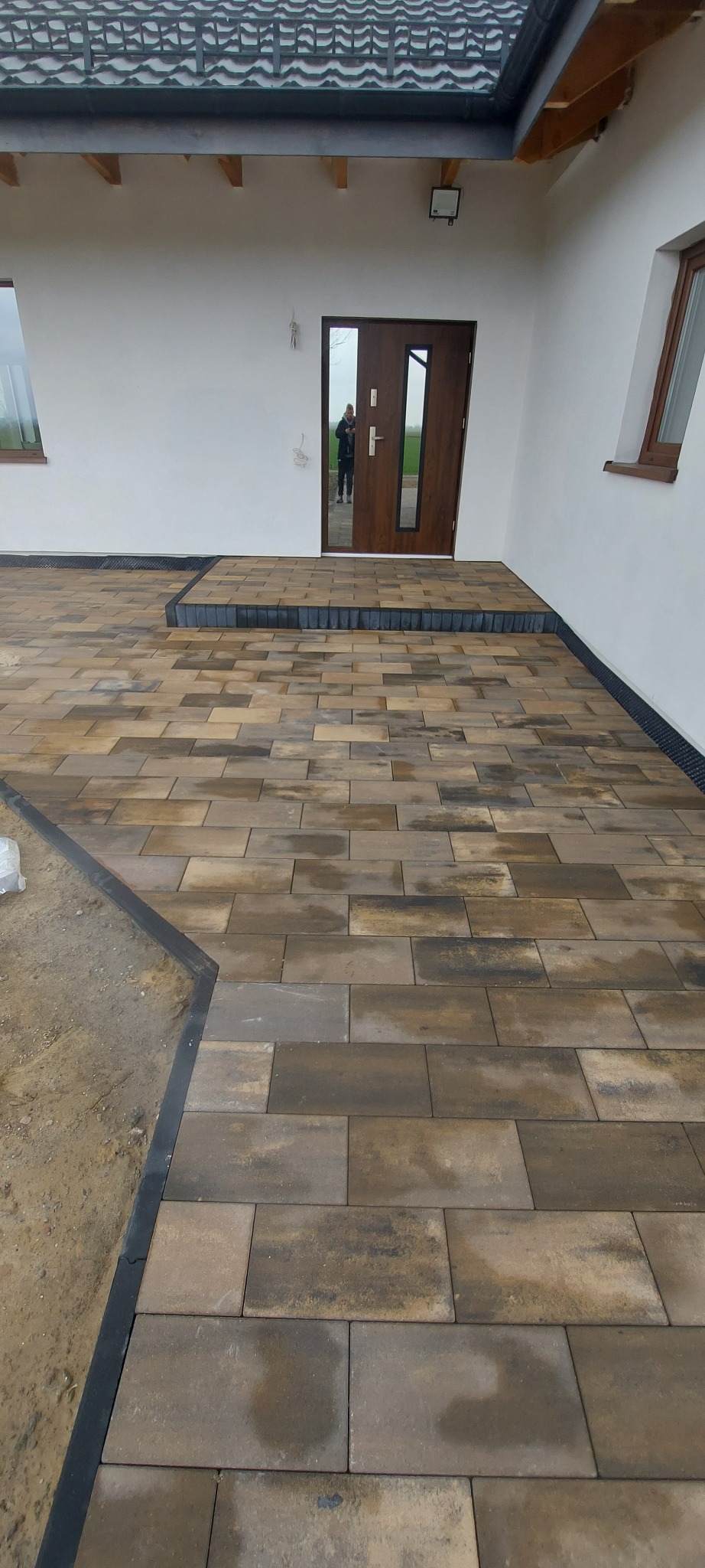 Ułożona kostka brukowa w odcieniach brązu i szarości, prowadząca do drzwi wejściowych z widocznym odbiciem osoby. Obrzeża wykończone czarnym elementem.
