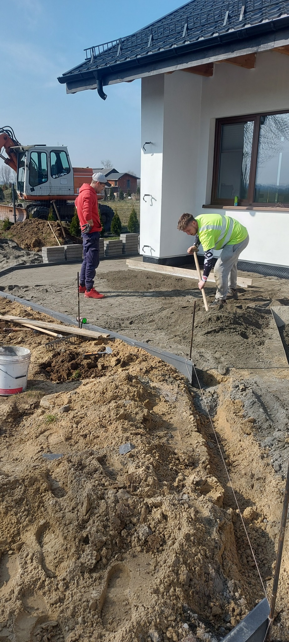 Prace brukarskie przy domu: robotnik wyrównuje podłoże pod kostkę, drugi nadzoruje. W tle koparka i ułożone kostki. Widoczne narzędzia i przygotowany teren.