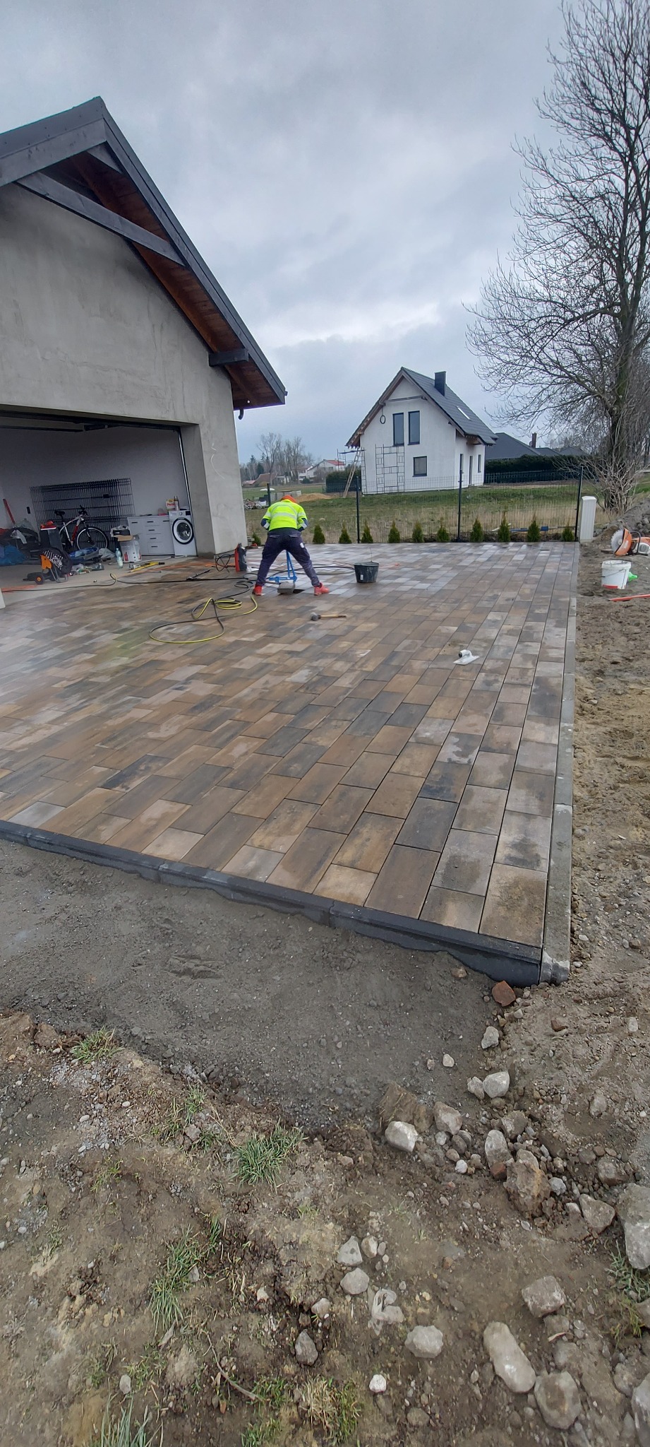 Pracownik w żółtej kamizelce układa kostkę brukową na posesji. Widoczny dom z garażem i drugi budynek w tle. Kostka ułożona na dużej powierzchni, obrzeża z czarnych elementów.