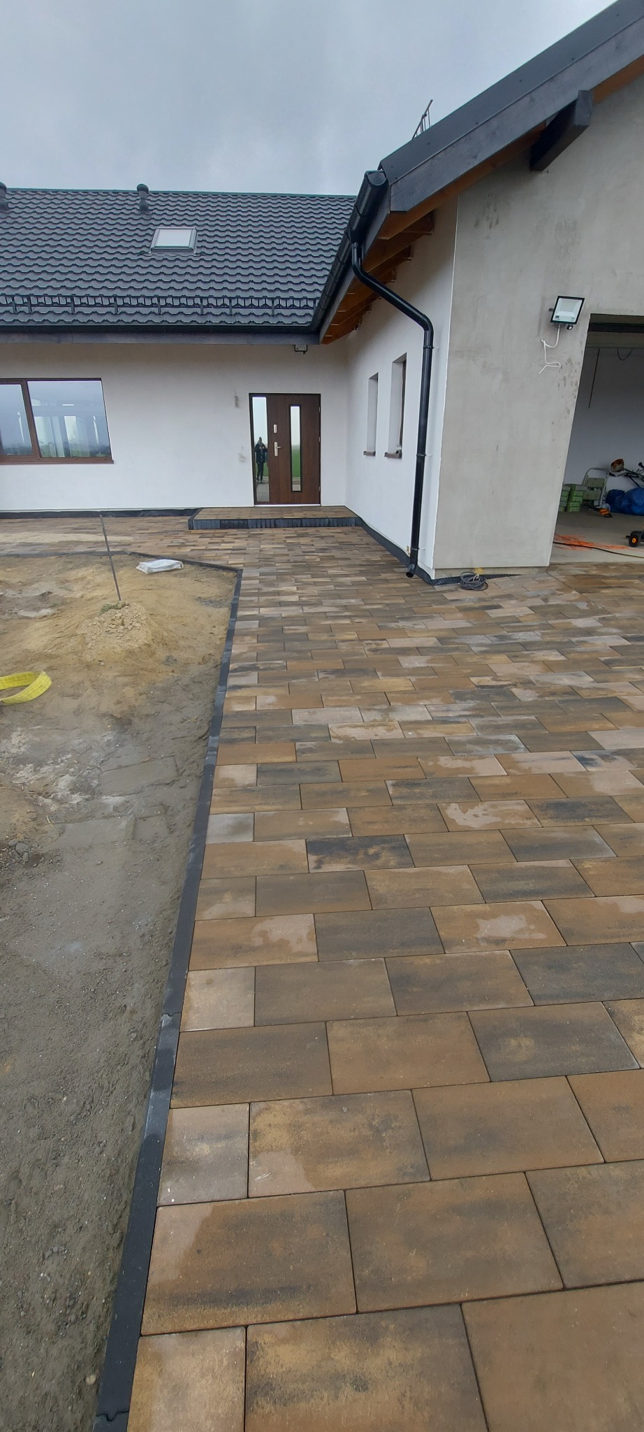 Ułożona kostka brukowa w odcieniach brązu przed domem z białymi ścianami i ciemnym dachem. Częściowo widoczne niezabrukowane podłoże.
