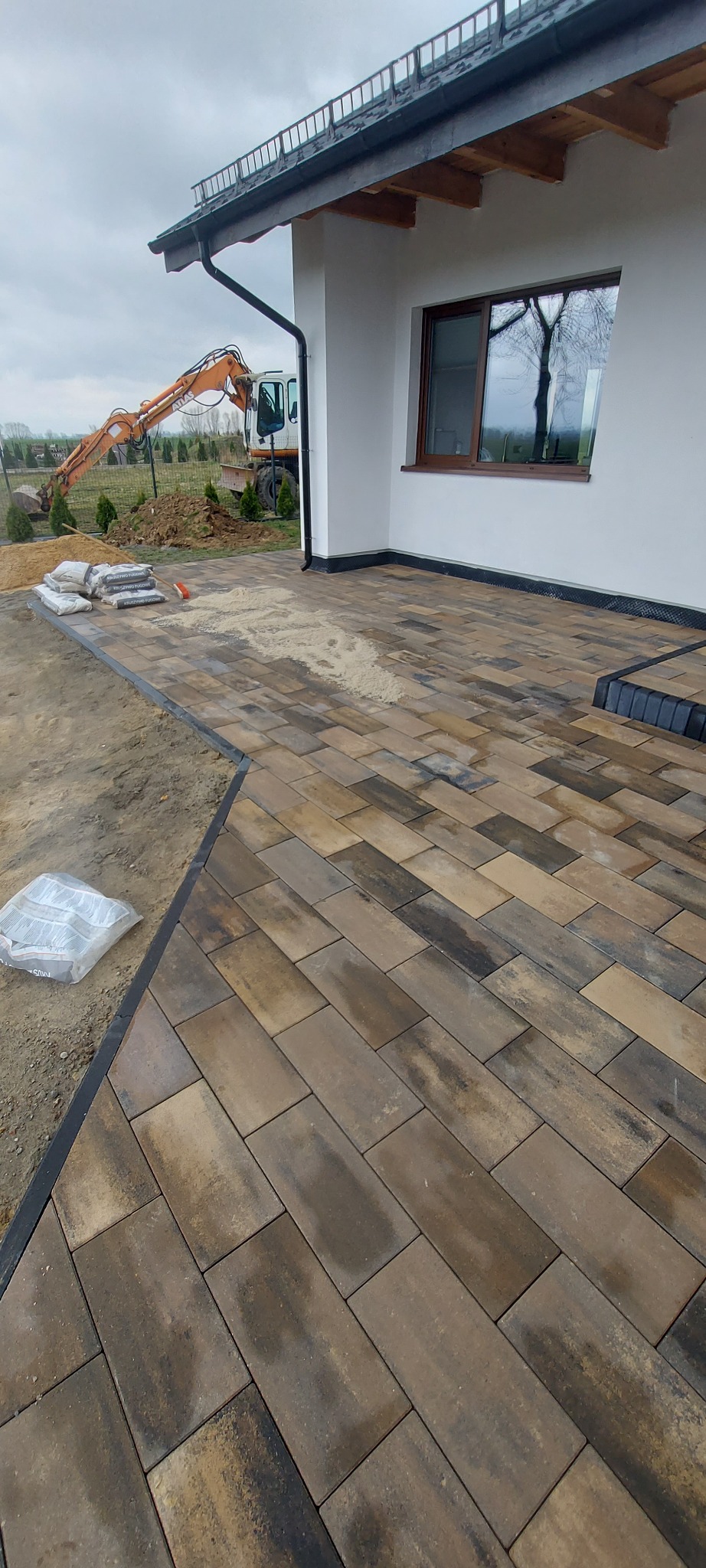 Ułożona kostka brukowa w odcieniach brązu przy budynku, z workami piasku i koparką w tle. Widoczny fragment tarasu i przygotowany teren pod dalsze prace.