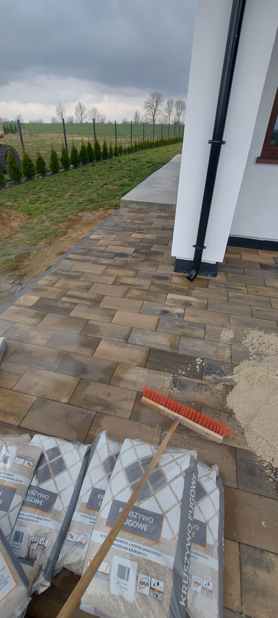 Ułożona kostka brukowa w odcieniach brązu, widoczne worki z kruszywem fugowym i szczotka do rozprowadzania, tło to trawnik i ogrodzenie.