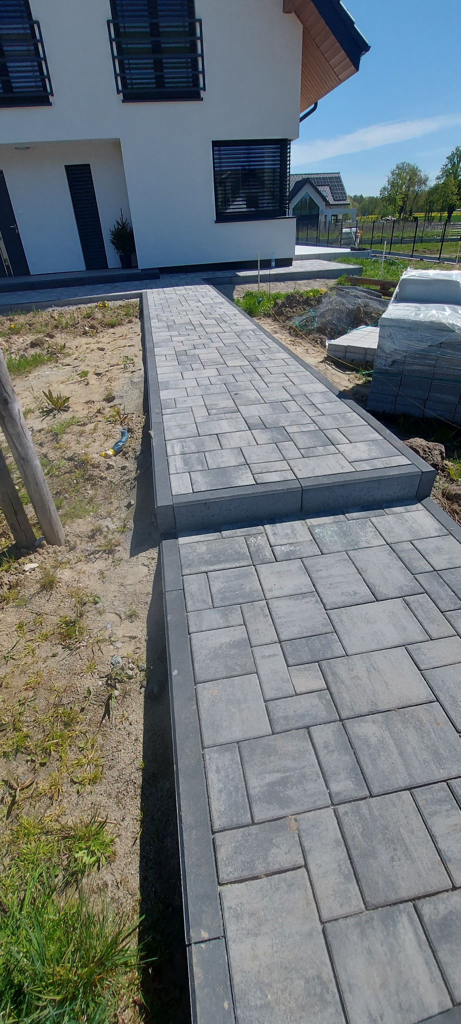Ułożona, szara kostka brukowa tworząca ścieżkę z obrzeżami na tle domu jednorodzinnego i sterty kostki. Widok z góry, słoneczny dzień.