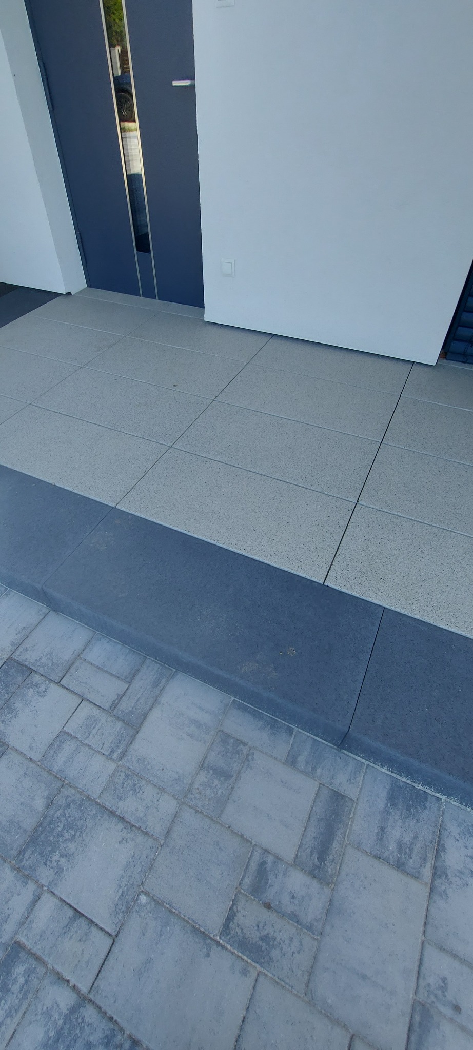 Nowoczesne wejście do domu z szarej kostki brukowej i dużych płyt, wykończone ciemnoszarym obrzeżem. Widoczne drzwi wejściowe i fragment białej ściany.