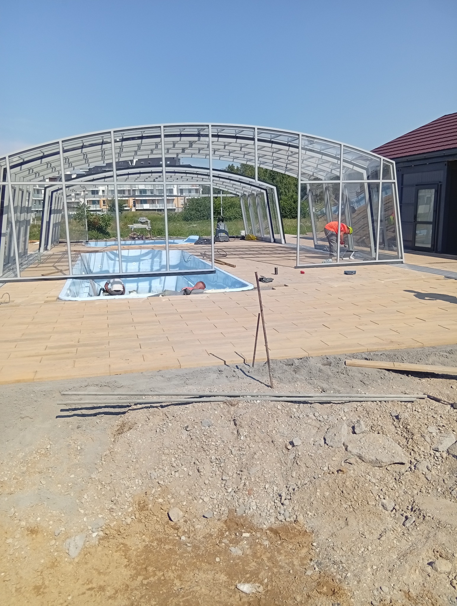 Montaż zadaszenia basenu z poliwęglanu na kostce. Pracownik w pomarańczowej koszulce przy pracy. Widok na budynek i zieleń w tle.